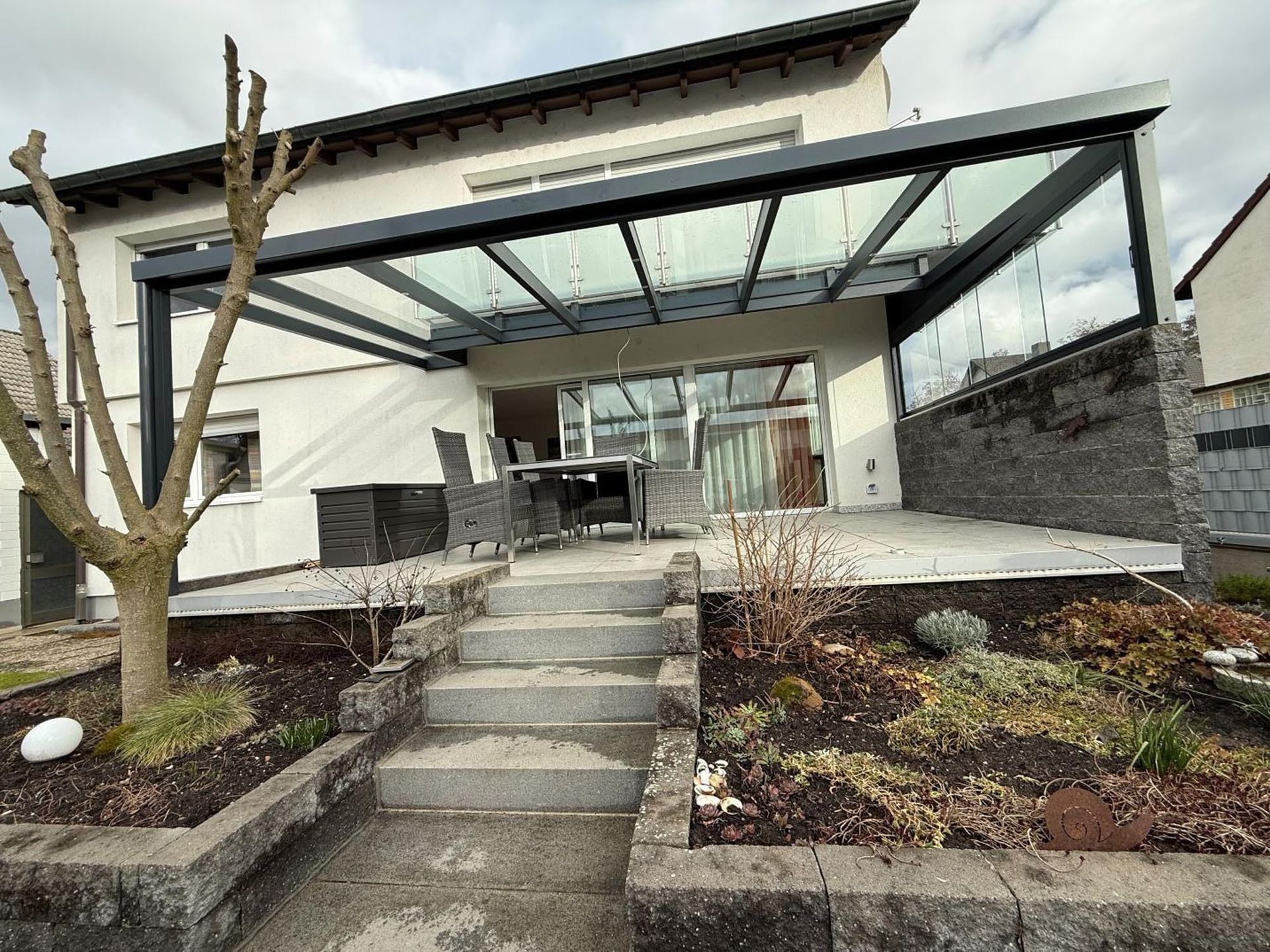 Moderner Hauseingang mit Betontreppe, Glasvordach und angelegtem Garten unter bewölktem Himmel