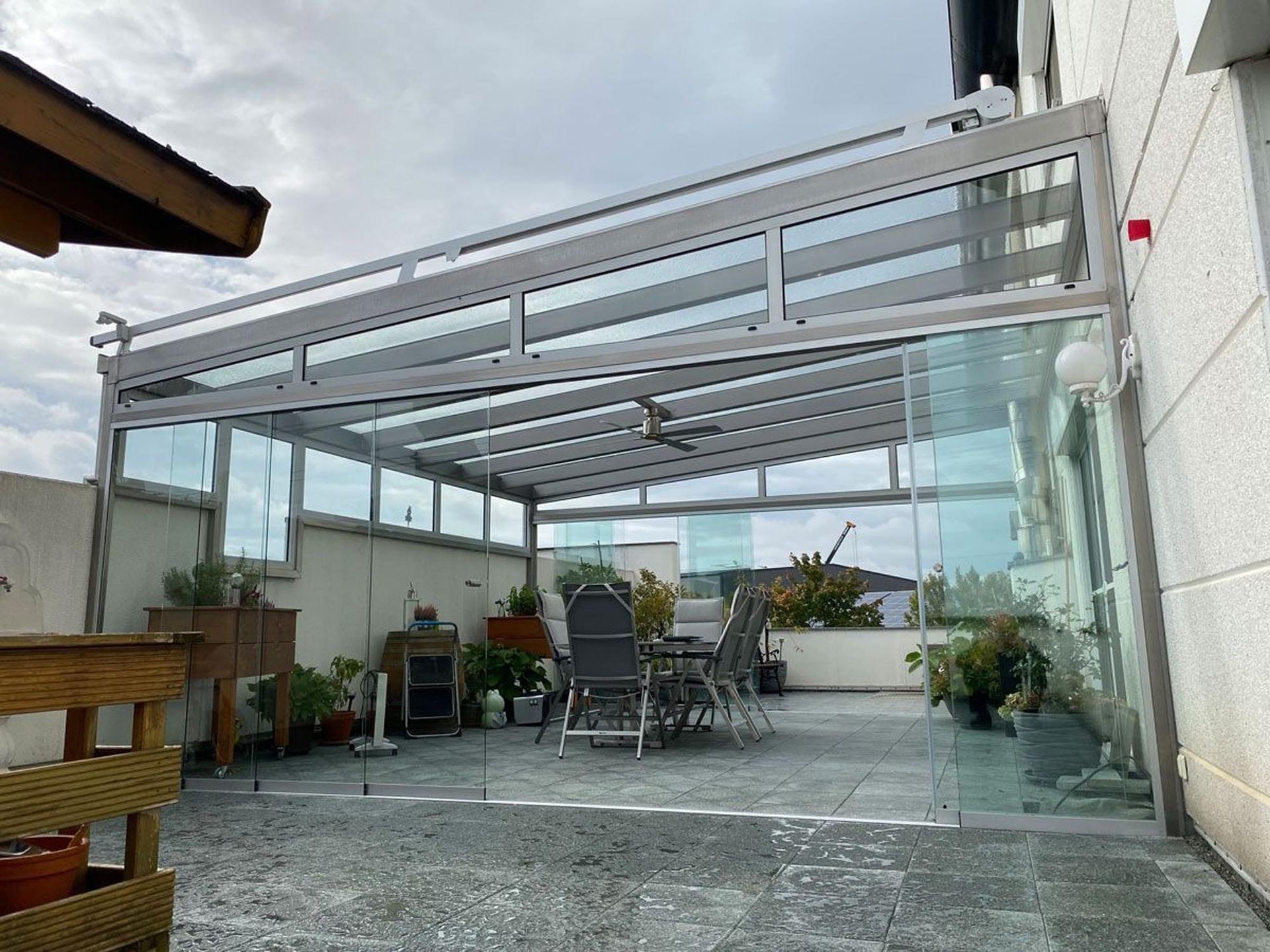 Überdachte Dachterrasse mit Glaswänden, grau gefliestem Boden und Esstisch im Freien unter einer weißen Pergola.