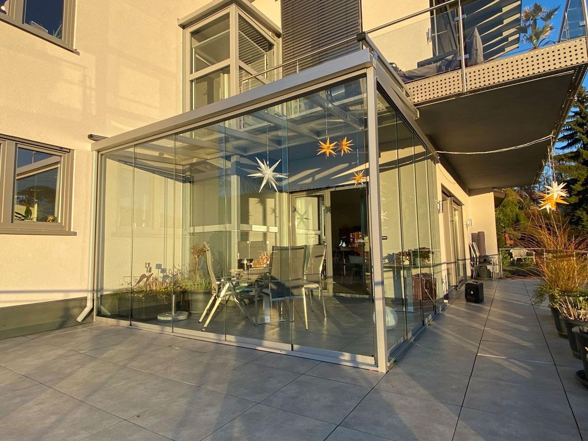 Glasumrandete Café-Terrasse vor einem cremefarbenen Gebäude, mit Tischen und Stühlen im Freien im Sonnenlicht.