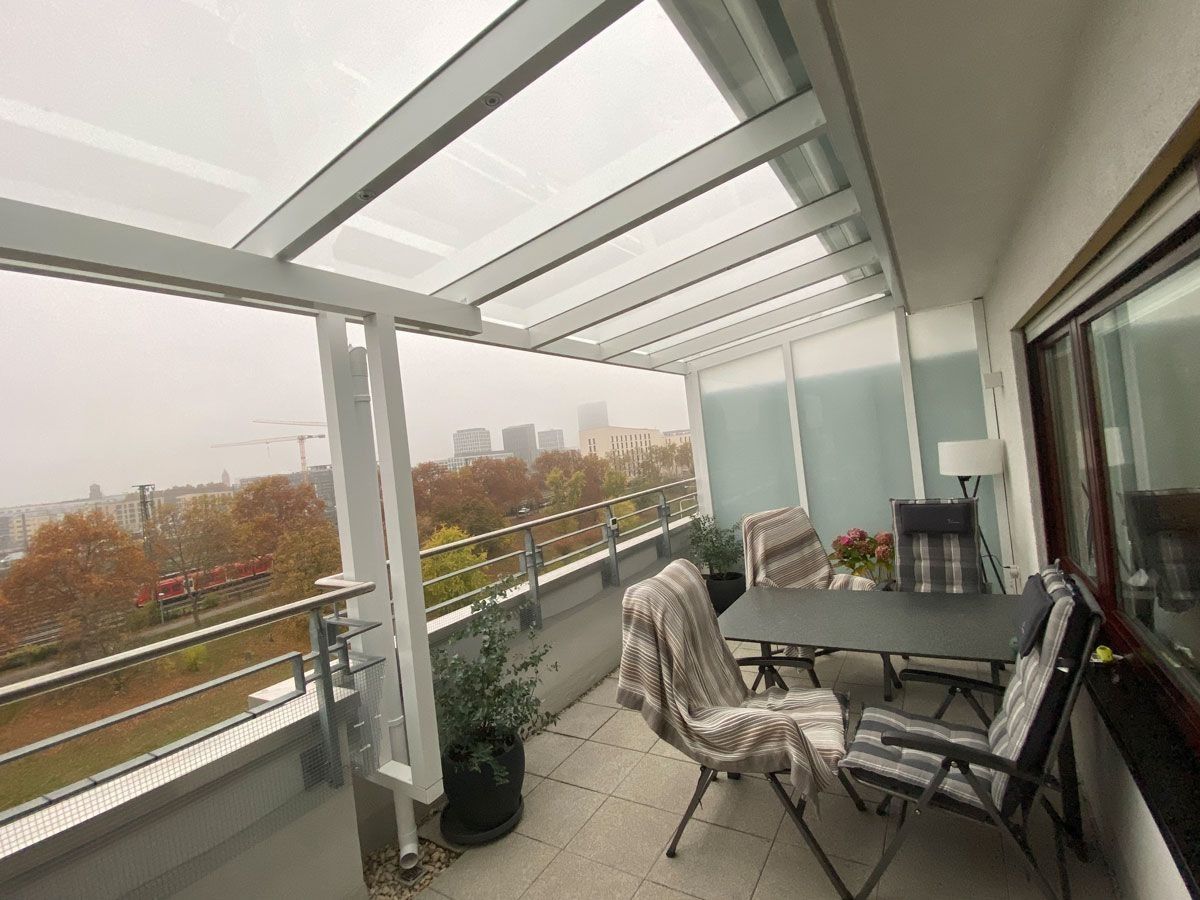 Überdachter Balkon mit Glasgeländer, Terrassentisch und Stühlen sowie Herbstbäumen im Außenbereich.