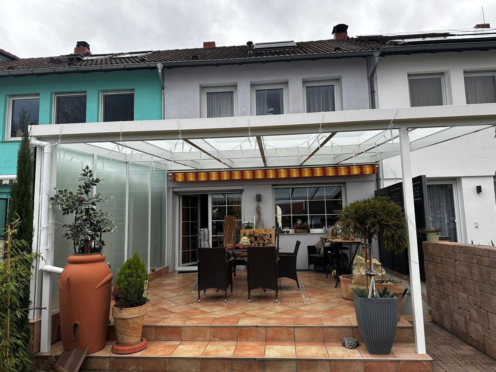 Terrasse mit Glasdach, Topfpflanzen, Esstisch und Stühlen vor einer Häuserreihe