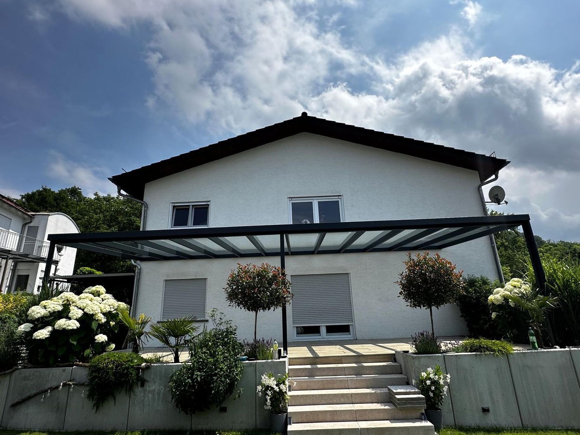 Weißes zweistöckiges Haus mit Veranda, Treppe und blühenden Sträuchern unter einem bewölkten Himmel