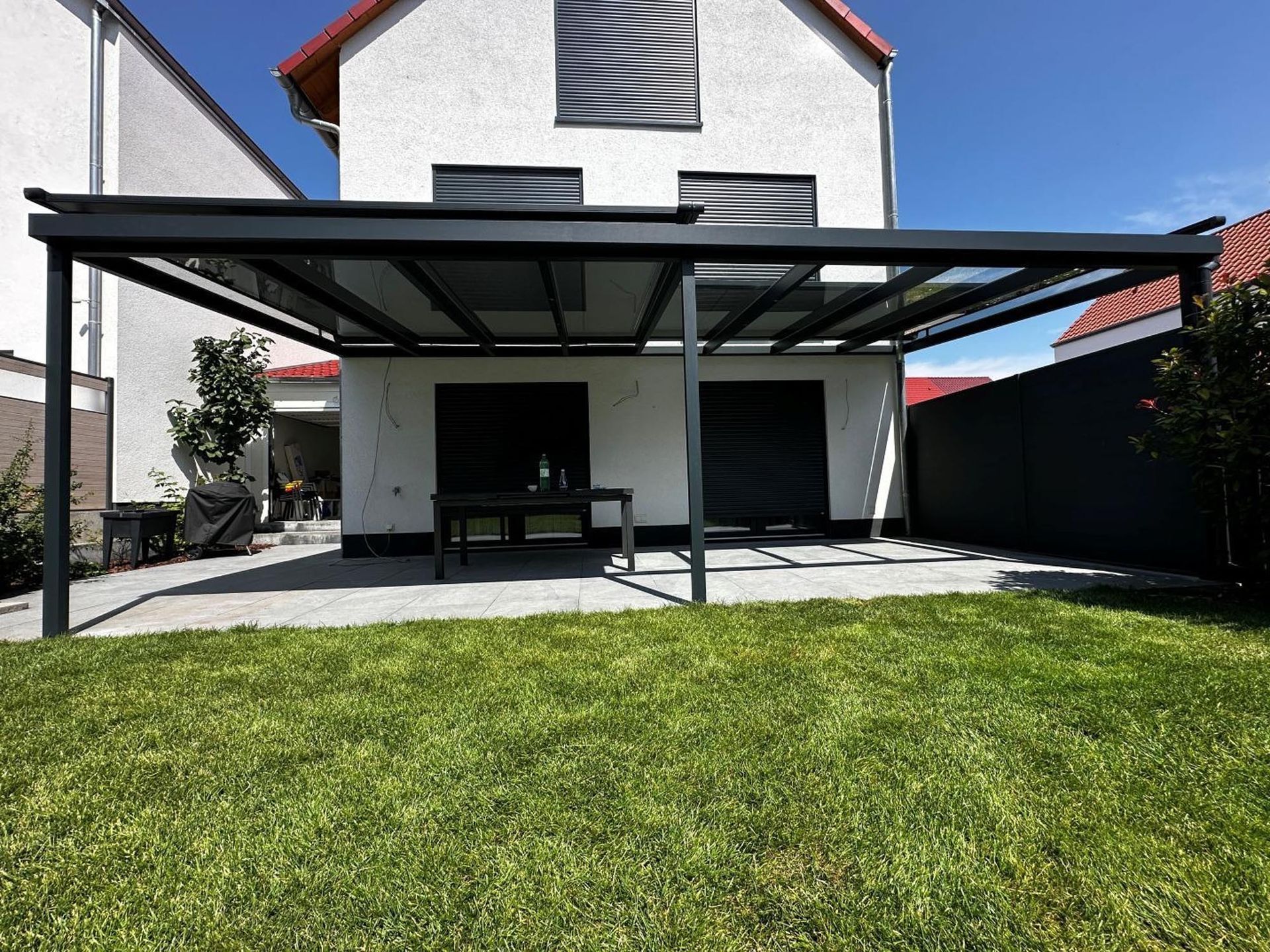 Hinterhofterrasse mit schwarzer Pergola, Bank und weißer Hauswand hinter einem grünen Rasen.