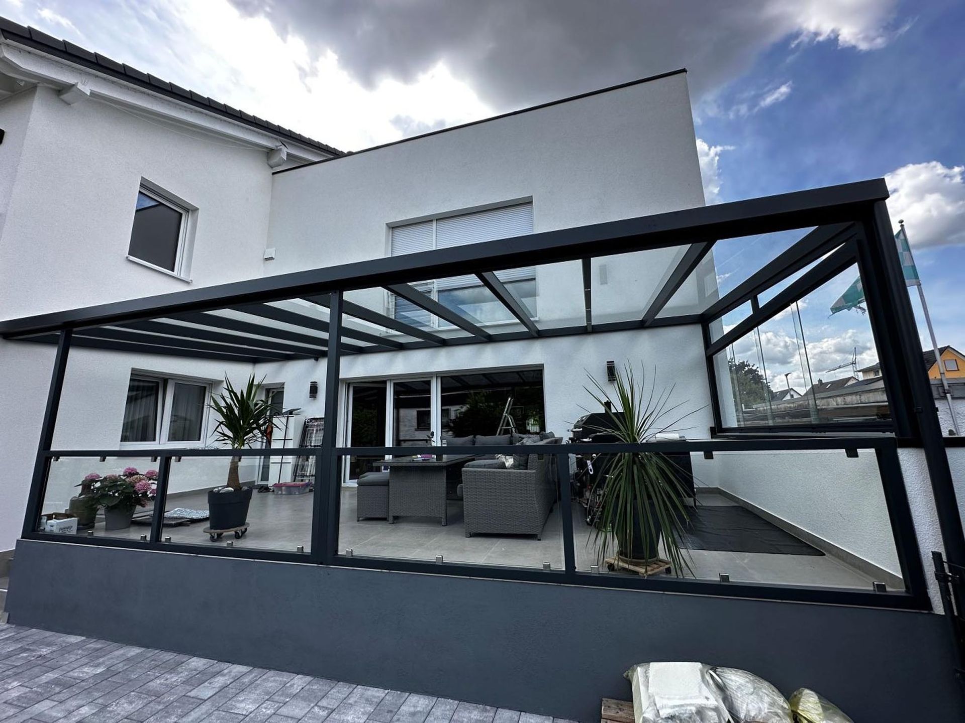 Moderne Terrasse mit Glasdachpergola an einem weißen Haus unter bewölktem Himmel