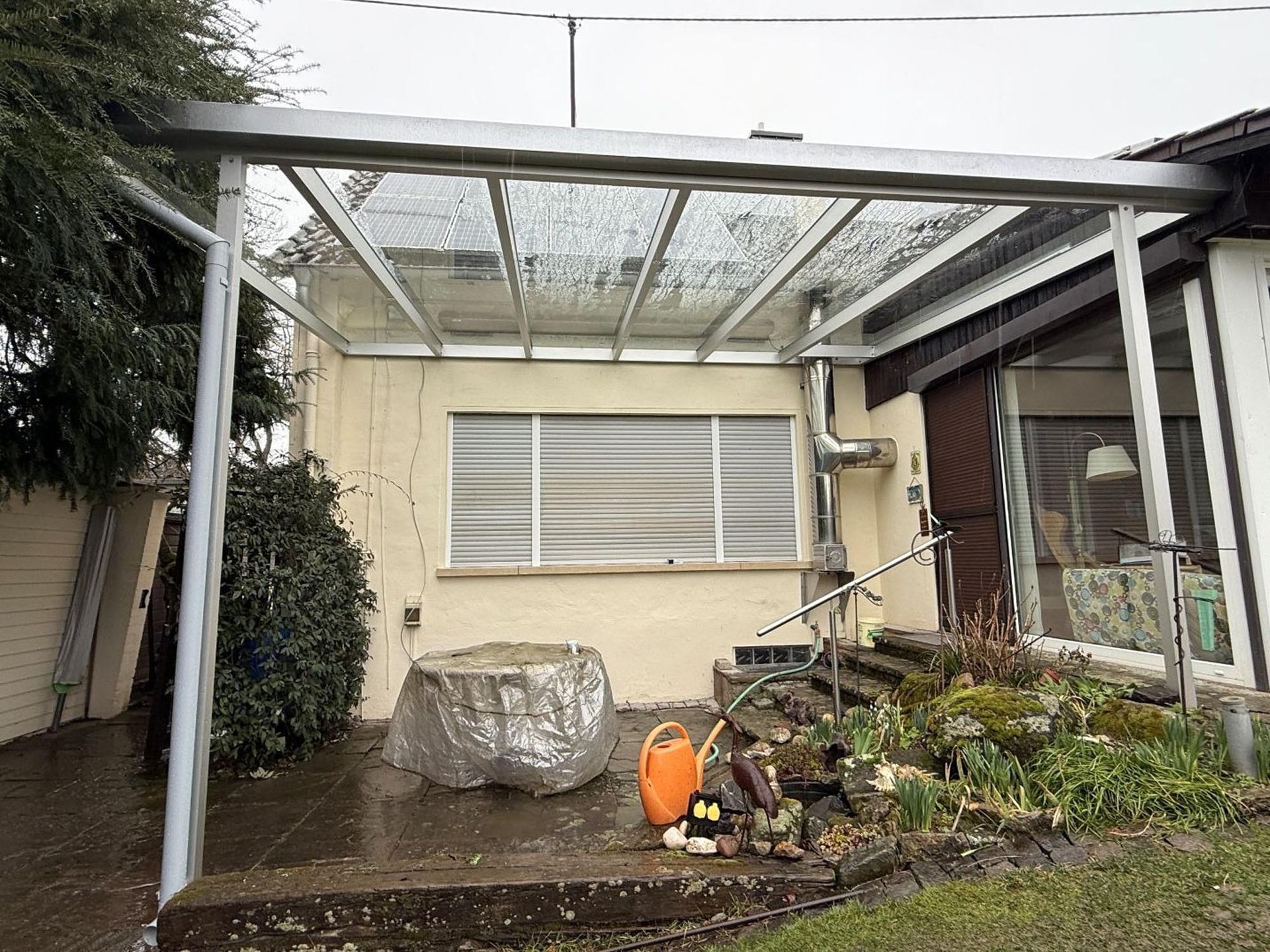 Hinterhofterrasse mit weißer Pergola, beigefarbener Hauswand, überdachten Gartenmöbeln und Topfpflanzen.
