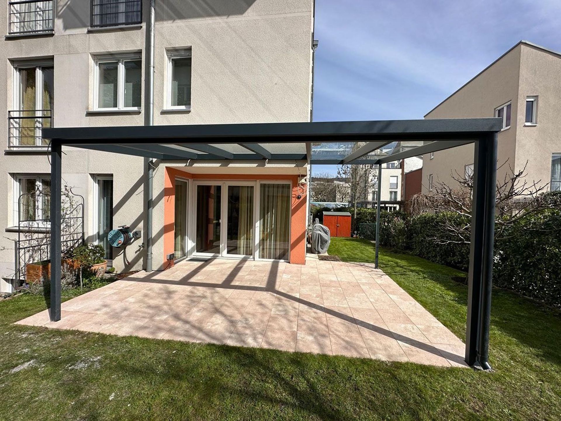 Moderne Terrasse mit schwarzer Pergola neben einem Haus, gefliester Terrasse, Rasen und Glasschiebetüren