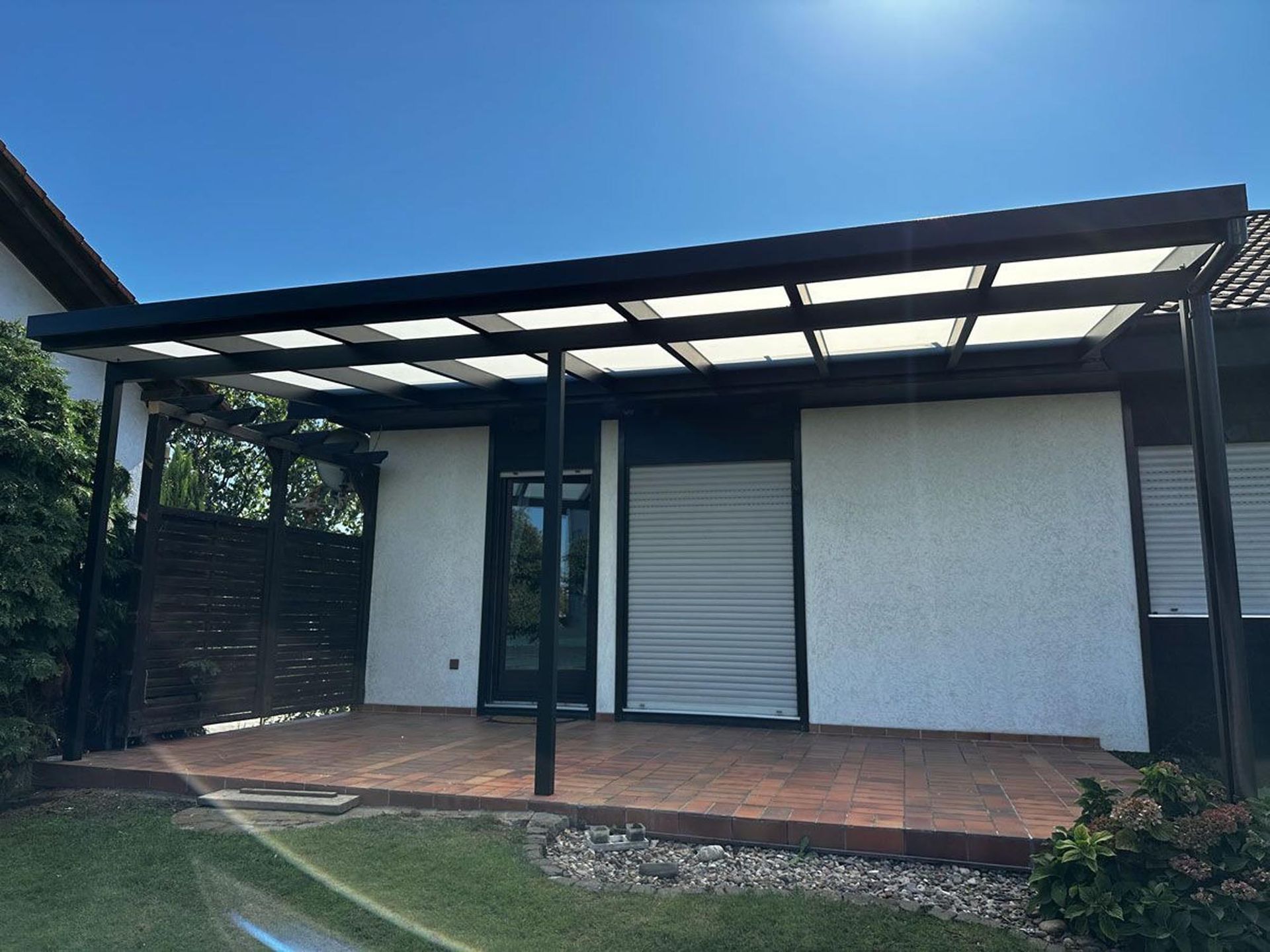 Moderne Hausterrasse mit schwarzer Metallpergola, weißen Wänden und Ziegelboden unter strahlend blauem Himmel