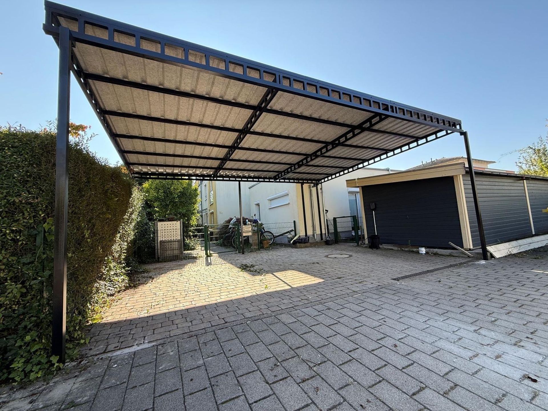Überdachte Auffahrt mit großem Carport aus Metall, gepflasterter Innenhof und Garagentore unter klarem Himmel
