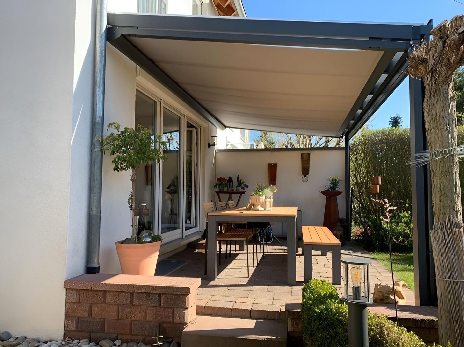Terrasse mit überdachter Pergola, Esstisch, Bänken, Topfpflanzen und einer gemauerten Stufe neben den Schiebetüren