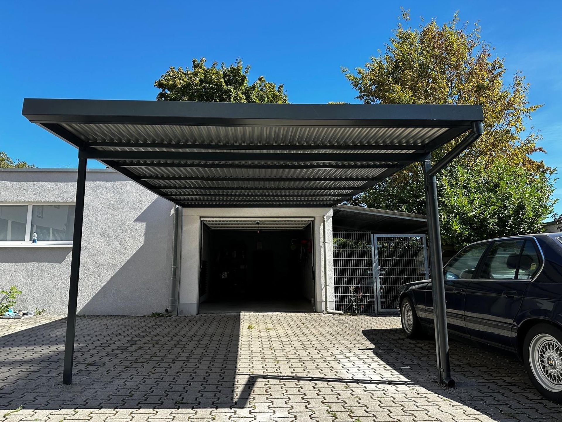 Überdachte Auffahrt mit schwarzem Metallcarport, offener Garage und geparktem SUV neben einem Haus.