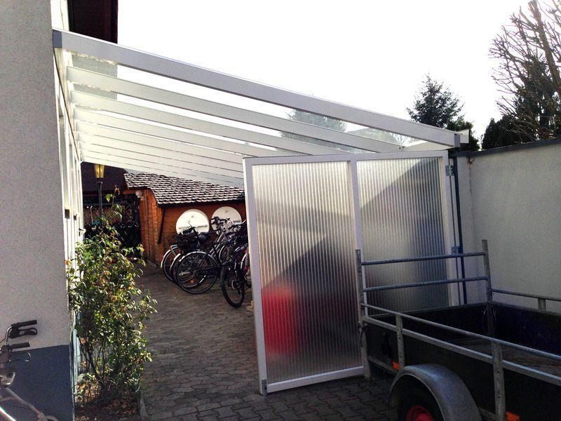 Weißer Carport mit mattiertem Schiebetor, geparkte Fahrräder und ein Anhänger in der Einfahrt