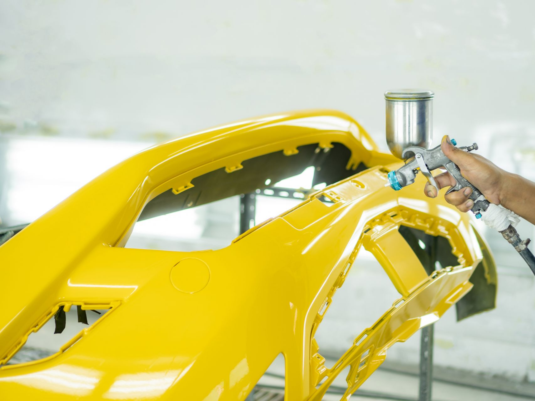 Une personne peint à la bombe un pare-chocs de voiture jaune dans un atelier.