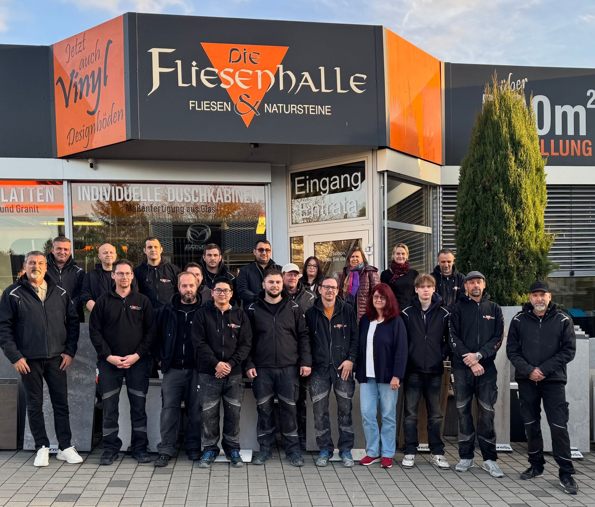 Eine Gruppe von Menschen steht vor einem Geschäft, „Die FliesenHalle“. Sie tragen schwarze Jacken.