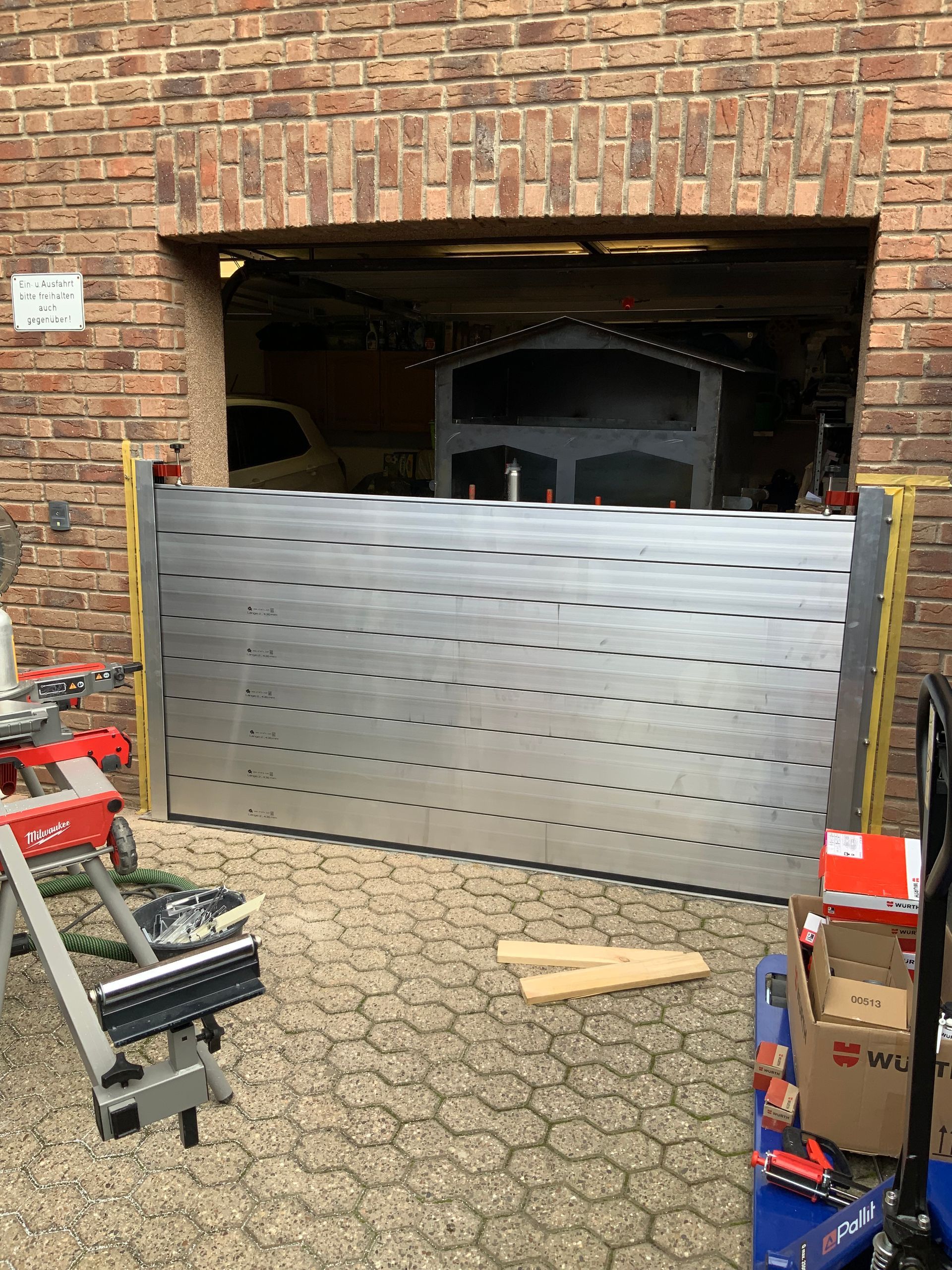 Montage eines Garagentors im Gange: silbernes Metalltor, Backsteingebäude, Werkzeuge und Pflastersteine.