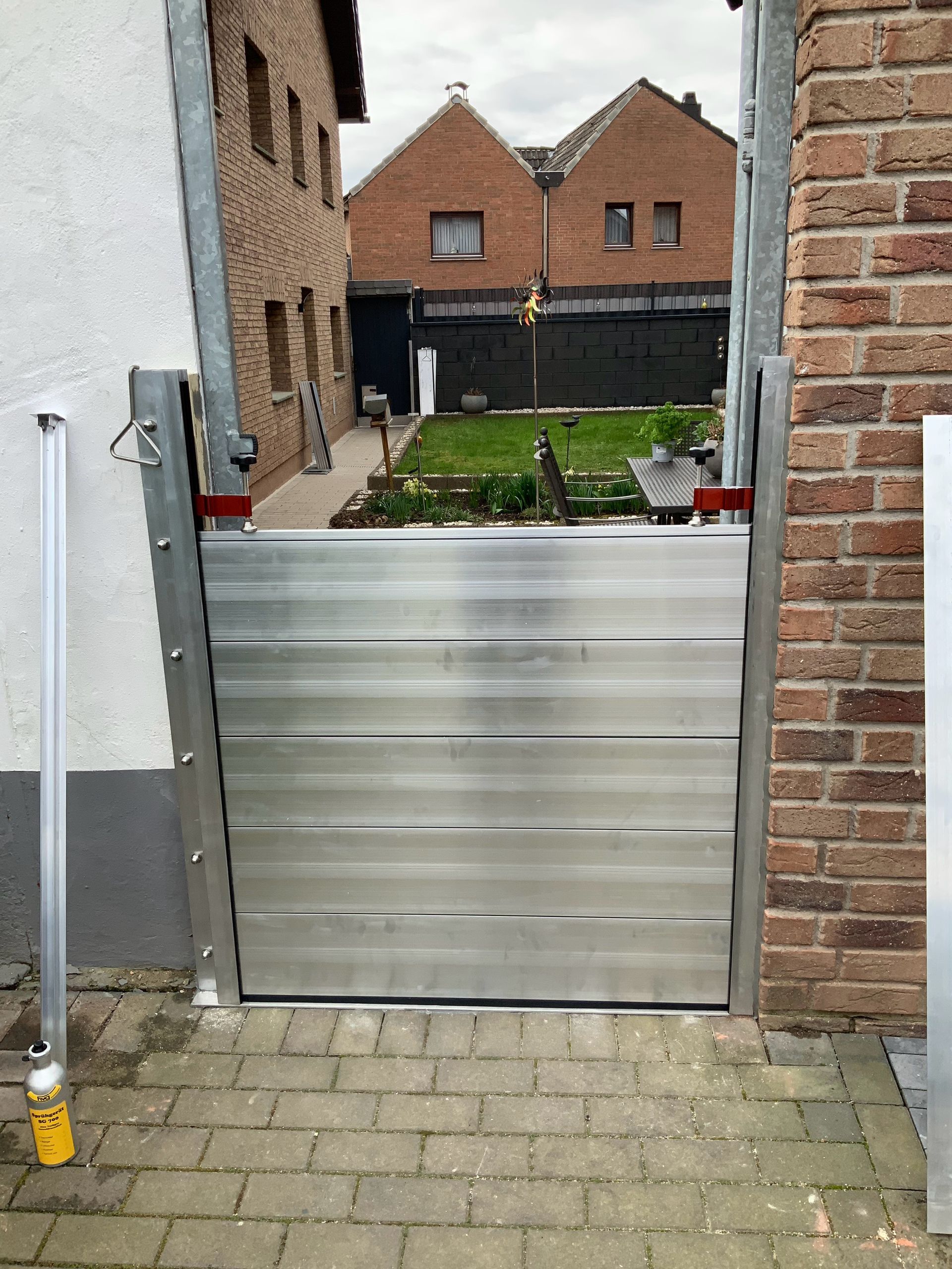 Eine Hochwasserschutzbarriere wurde in einem Türrahmen installiert und schützt einen Hof, im Hintergrund sind Häuser zu sehen.