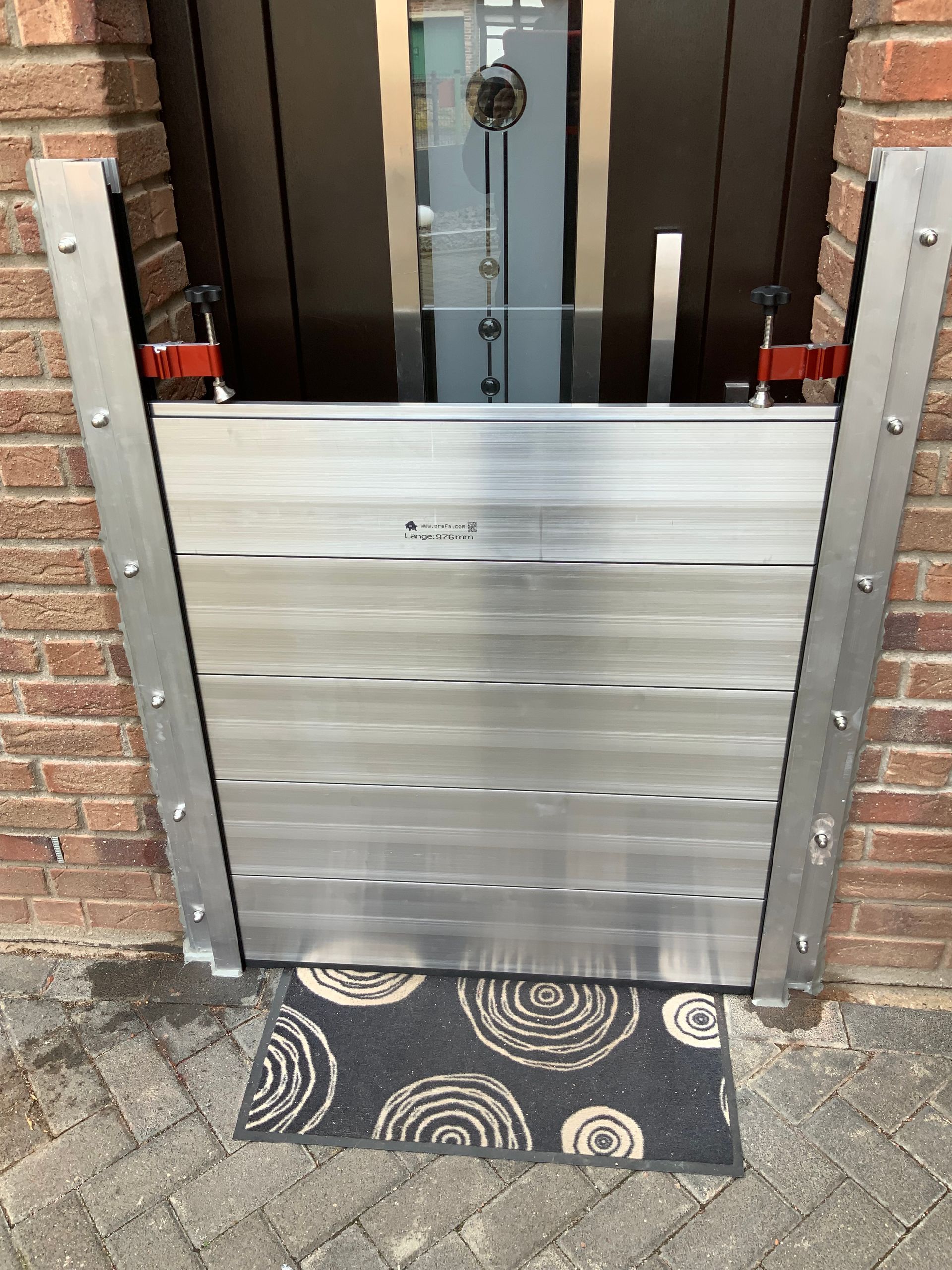 Eine Hochwasserschutzbarriere wurde vor einer Glastür installiert und mit Klemmen an einer Ziegelmauer befestigt.