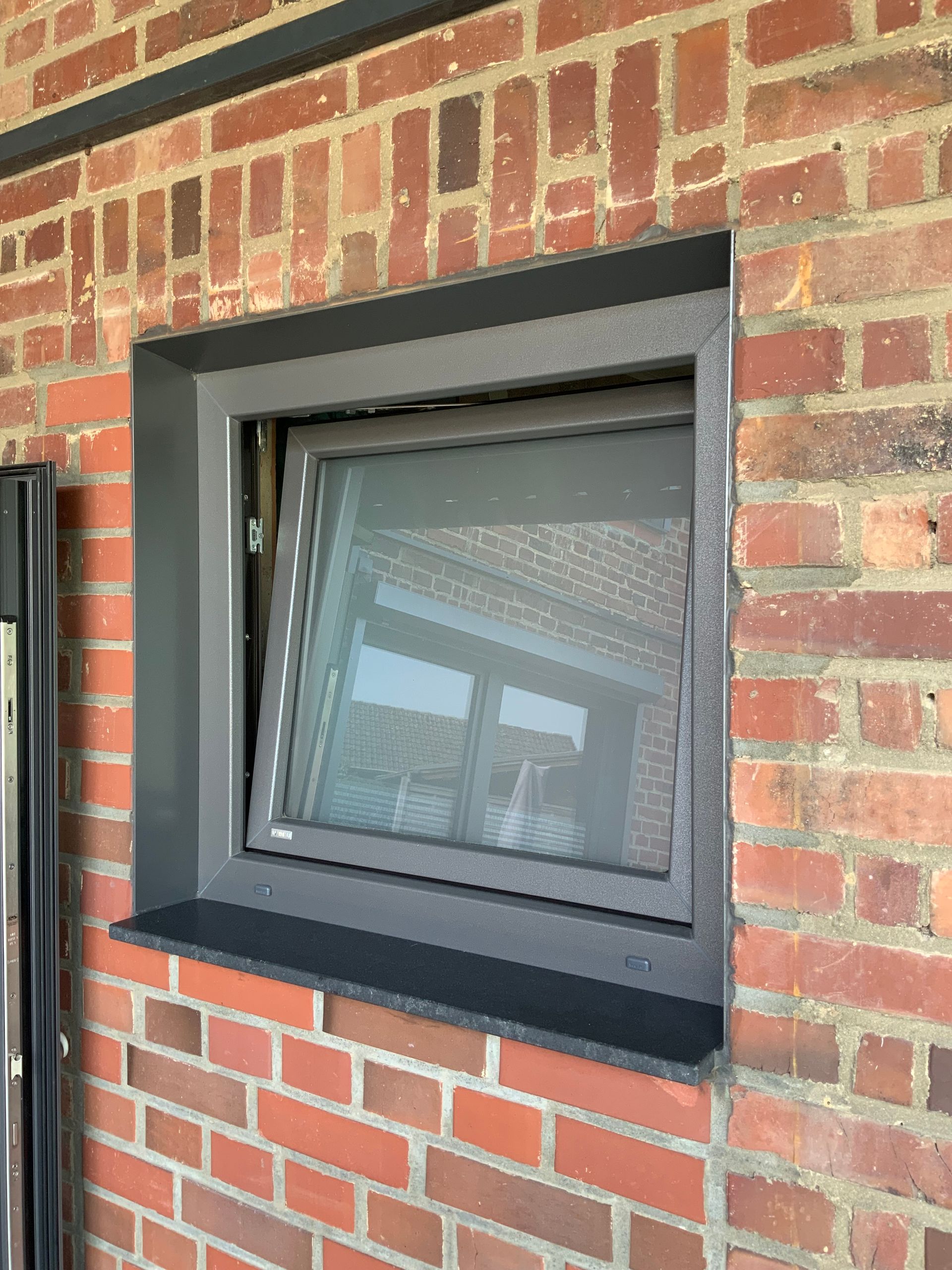 Ein dunkel gerahmtes Fenster in einer roten Backsteinmauer, leicht geöffnet. Unterhalb des Fensters ein dunkler Sims.