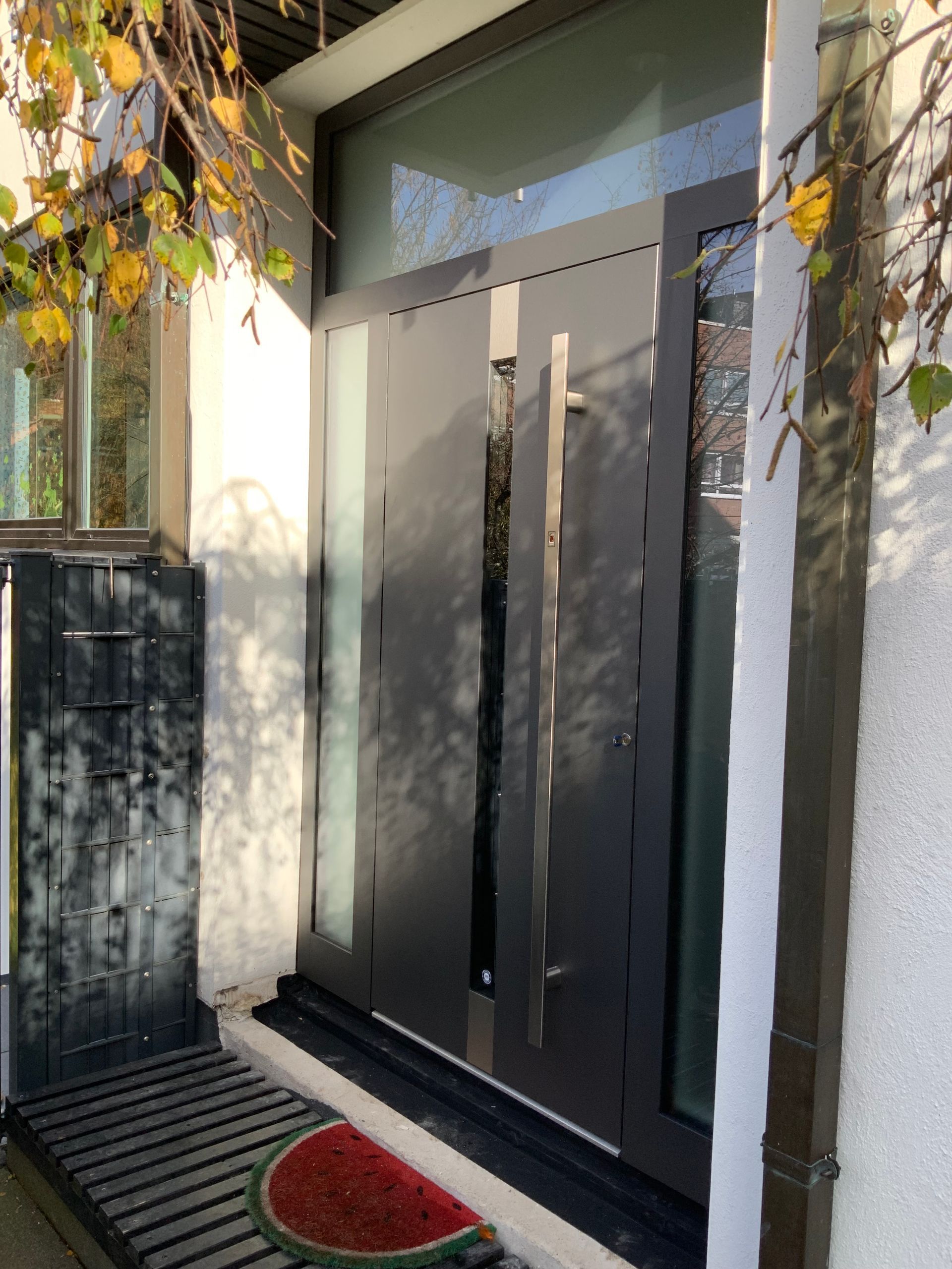 Graue Haustür mit langem Türgriff, seitlicher Glasscheibe und roter Fußmatte.