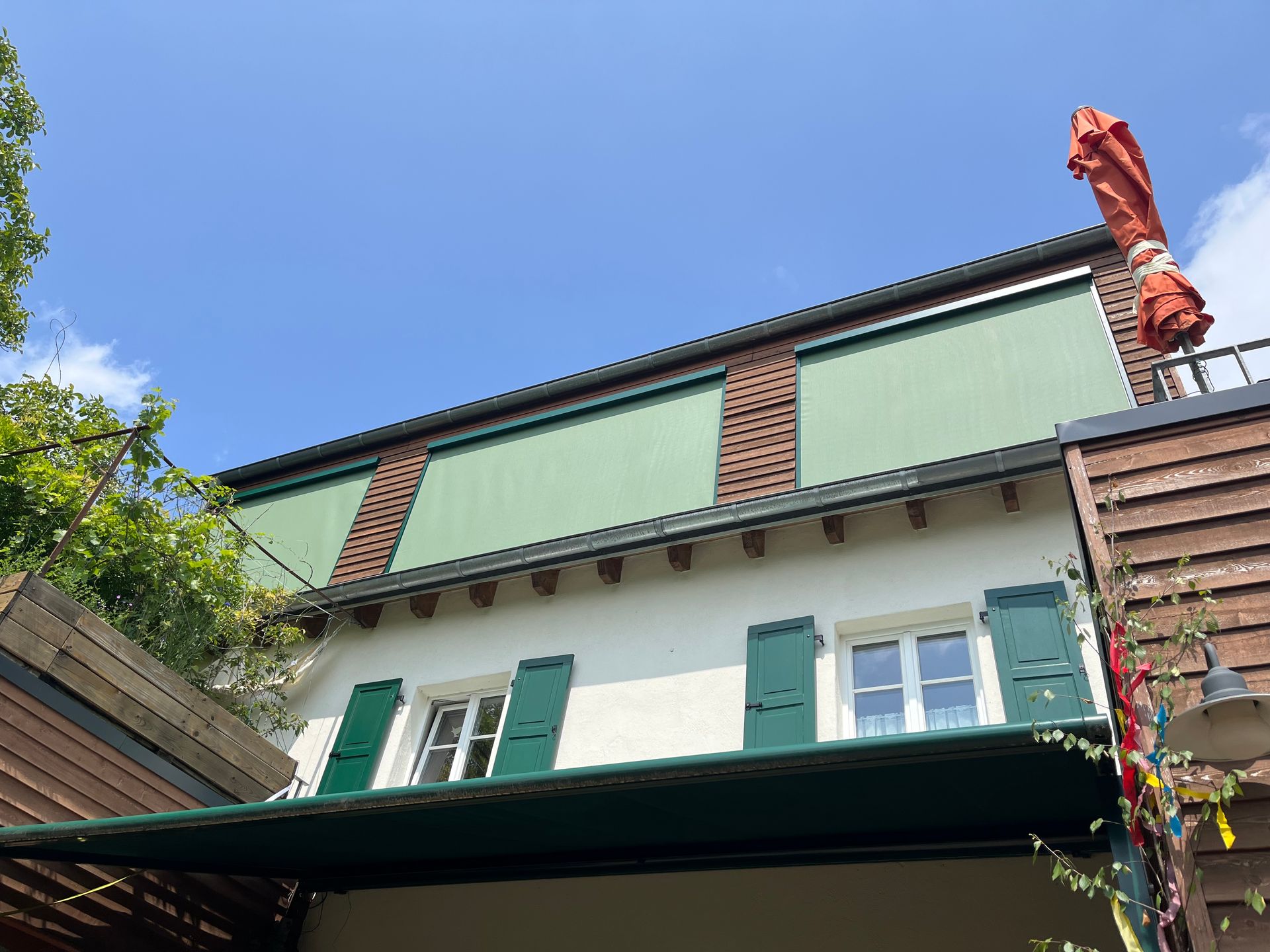 Zweistöckiges Gebäude mit grünen Fensterläden und Markise unter blauem Himmel, Backsteinakzenten und einem zusammengerollten roten Regenschirm.