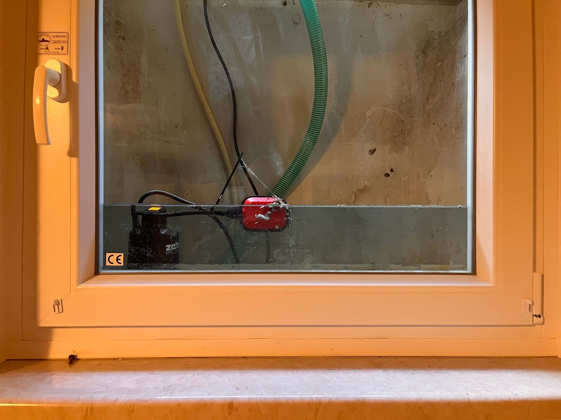 Eine Tauchpumpe im Inneren eines Fensters, teilweise unter Wasser, mit angeschlossenen Schläuchen und Stromkabeln.