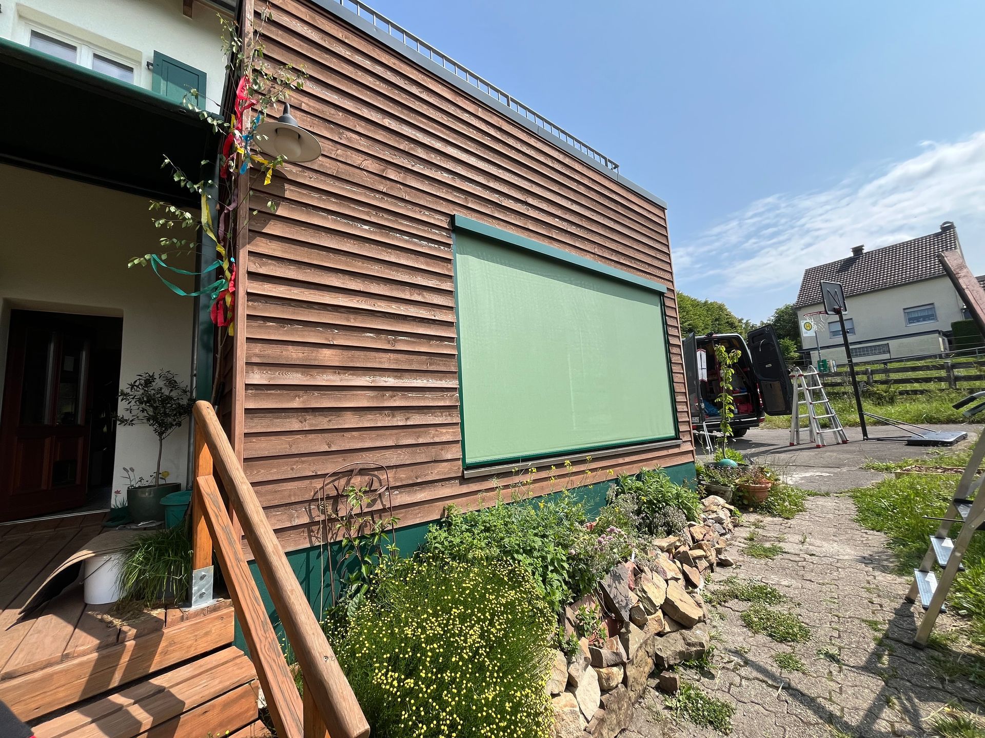 Holzgebäude mit grüner Markise. Im Vordergrund befinden sich eine Steineinfassung und Pflanzen.