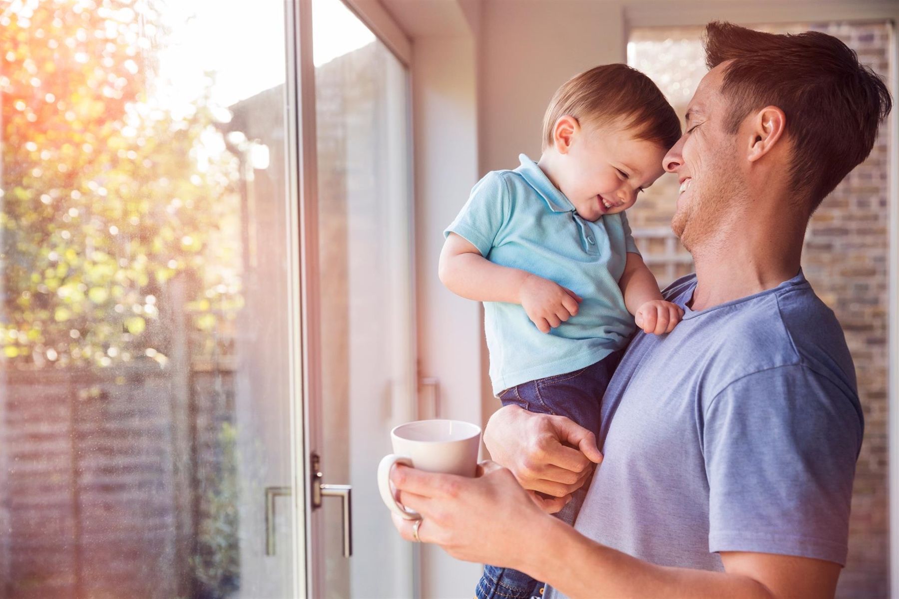 Vater hält ein lachendes Kleinkind an einem sonnigen Fenster, beide lächeln.