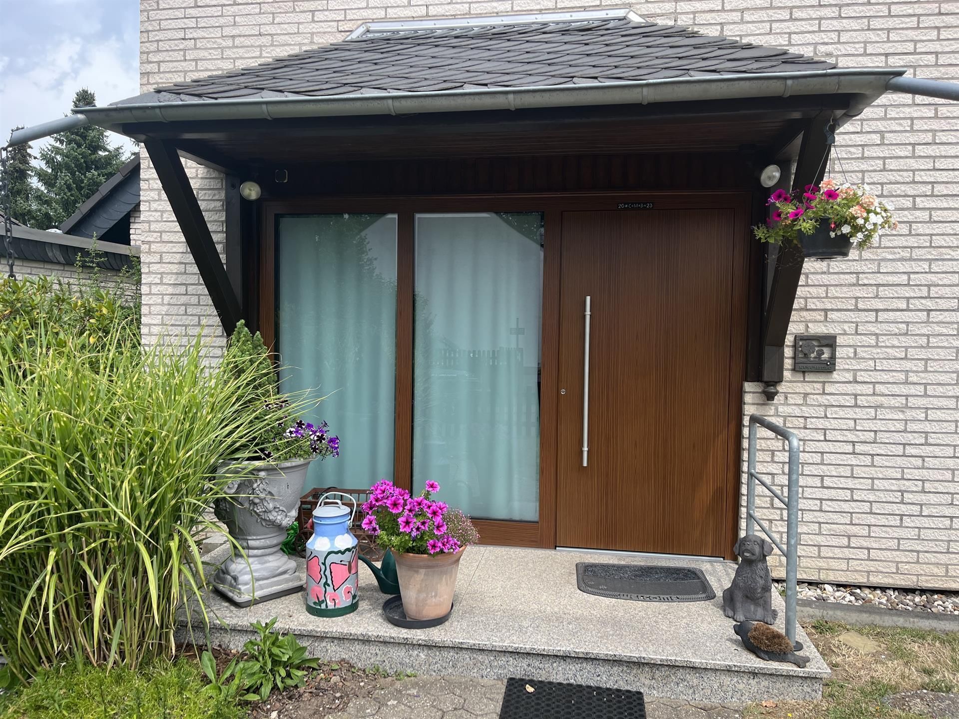 Braune Haustür mit Glasscheibe unter einem Holzvordach. Blumen und Pflanzen in Töpfen schmücken den Eingang.