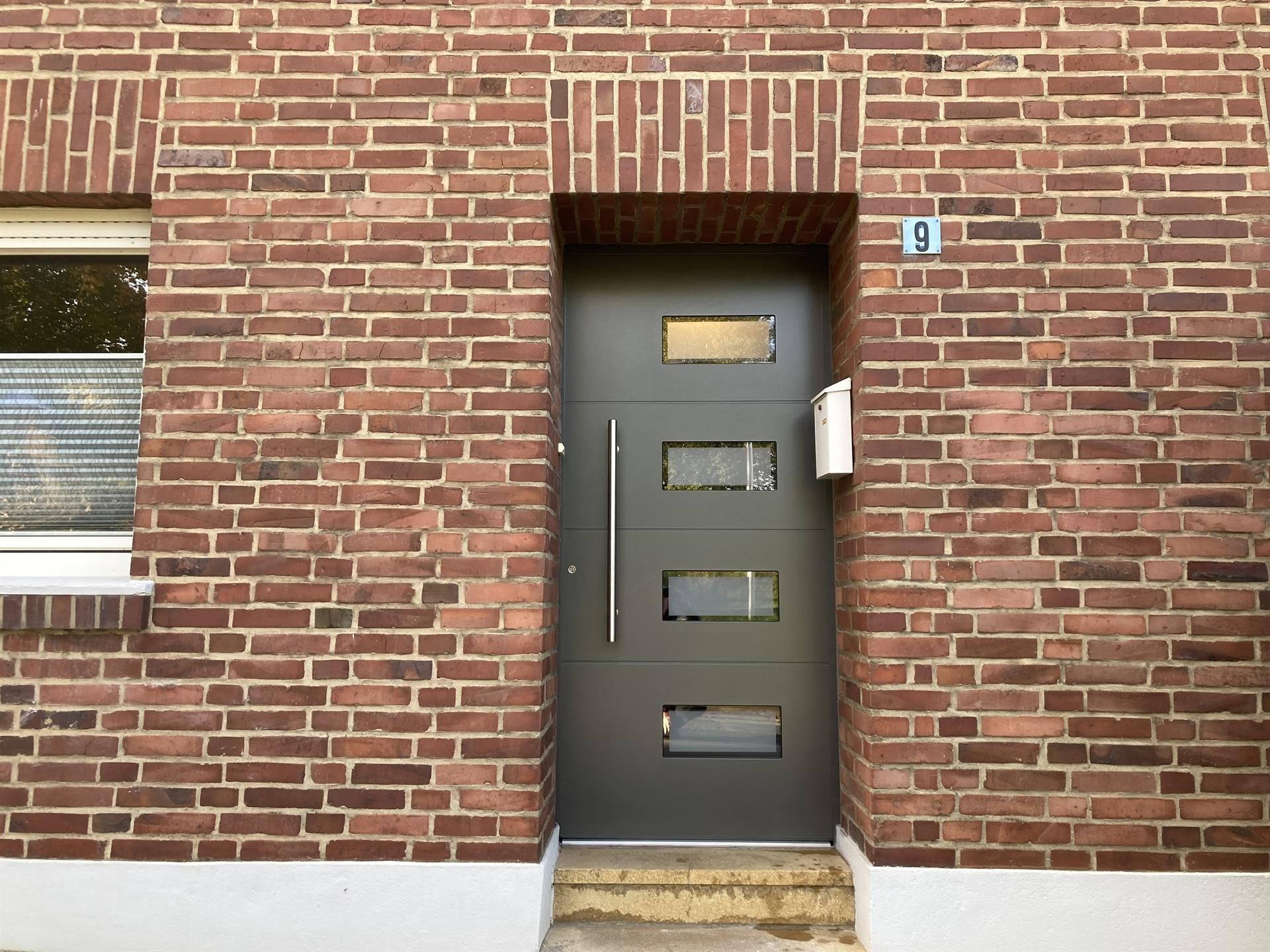 Graue Haustür mit rechteckigen Fenstern in einer Ziegelmauer. Die Tür hat einen silbernen Griff und befindet sich neben einem weißen Briefkasten.