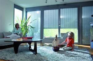 Frau auf der Couch, Kind spielt mit Spielzeugschiff auf Teppich im Wohnzimmer mit großen Fenstern und Jalousien.