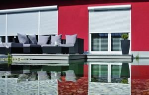 Rote Gebäudefassade mit weißen Rollläden, einem Sofa mit Kissen und einer Spiegelung im Wasser.