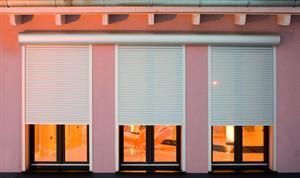 Drei Fenster mit weißen, geschlossenen Rollläden an einer rosafarbenen Gebäudefassade.