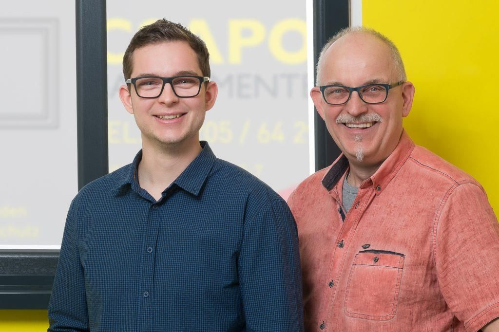 Zwei lächelnde Männer mit Brillen; einer in einem blauen Hemd, der andere in einem orangefarbenen Hemd, vor einer gelben Wand.