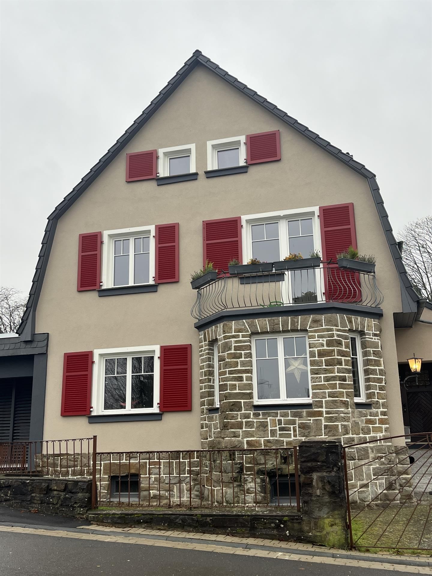 Hellbraunes Haus mit roten Fensterläden, Steinfassade, Balkon mit Pflanzgefäßen, bewölkter Himmel.