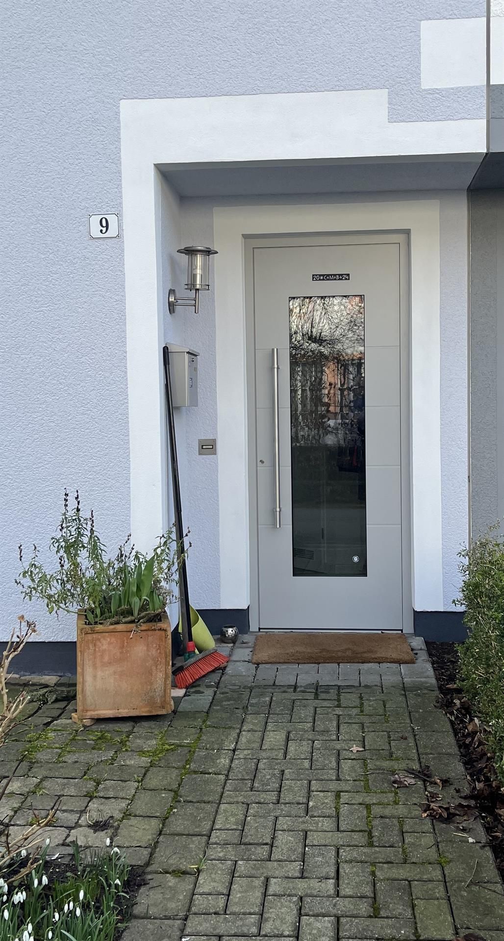 Graue Tür mit Glasscheibe, die zu einem Haus mit einem gemauerten Gehweg, einem Briefkasten und Topfpflanzen führt.