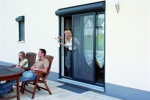Frau kommt mit Blumen in der Hand aus einer Glasschiebetür, ihre Familie sitzt an einem Tisch auf einer Terrasse.