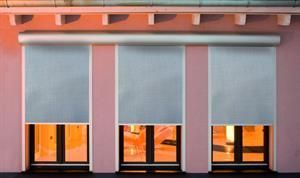 Drei helle Rollos verdecken teilweise die Fenster eines rosafarbenen Gebäudes.