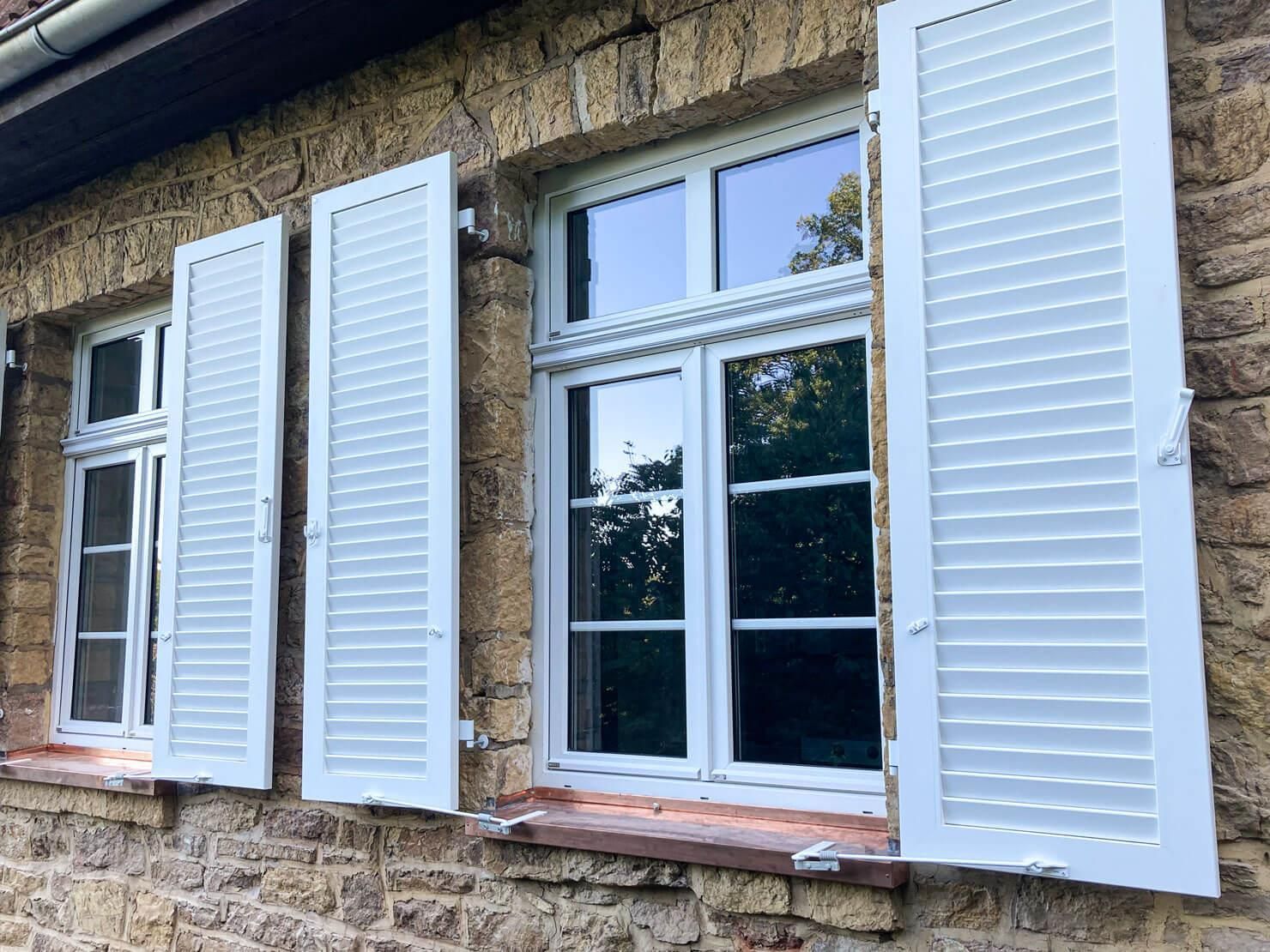 An den Fenstern eines Backsteingebäudes öffnen sich weiße Fensterläden.