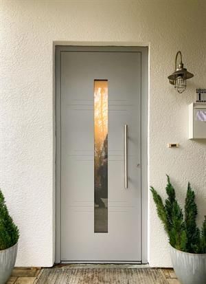 Graue Haustür mit vertikalem Fenster, Edelstahlgriff und Topfpflanzen.