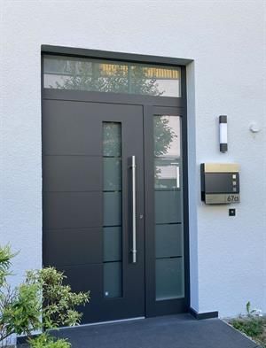 Dunkelgraue Haustür mit Glaseinsätzen und langem silbernen Griff, Briefkasten und Leuchte an weißer Wand.