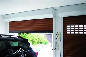 Braunes Rolltor der Garage teilweise geöffnet, Auto im Vordergrund. Eine weitere geschlossene Tür mit kleinem quadratischen Fenster.