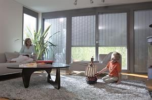 Frau auf der Couch, Kind mit Spielzeugschiff, große Fenster mit Jalousien, Wohnzimmer in neutralen Tönen.