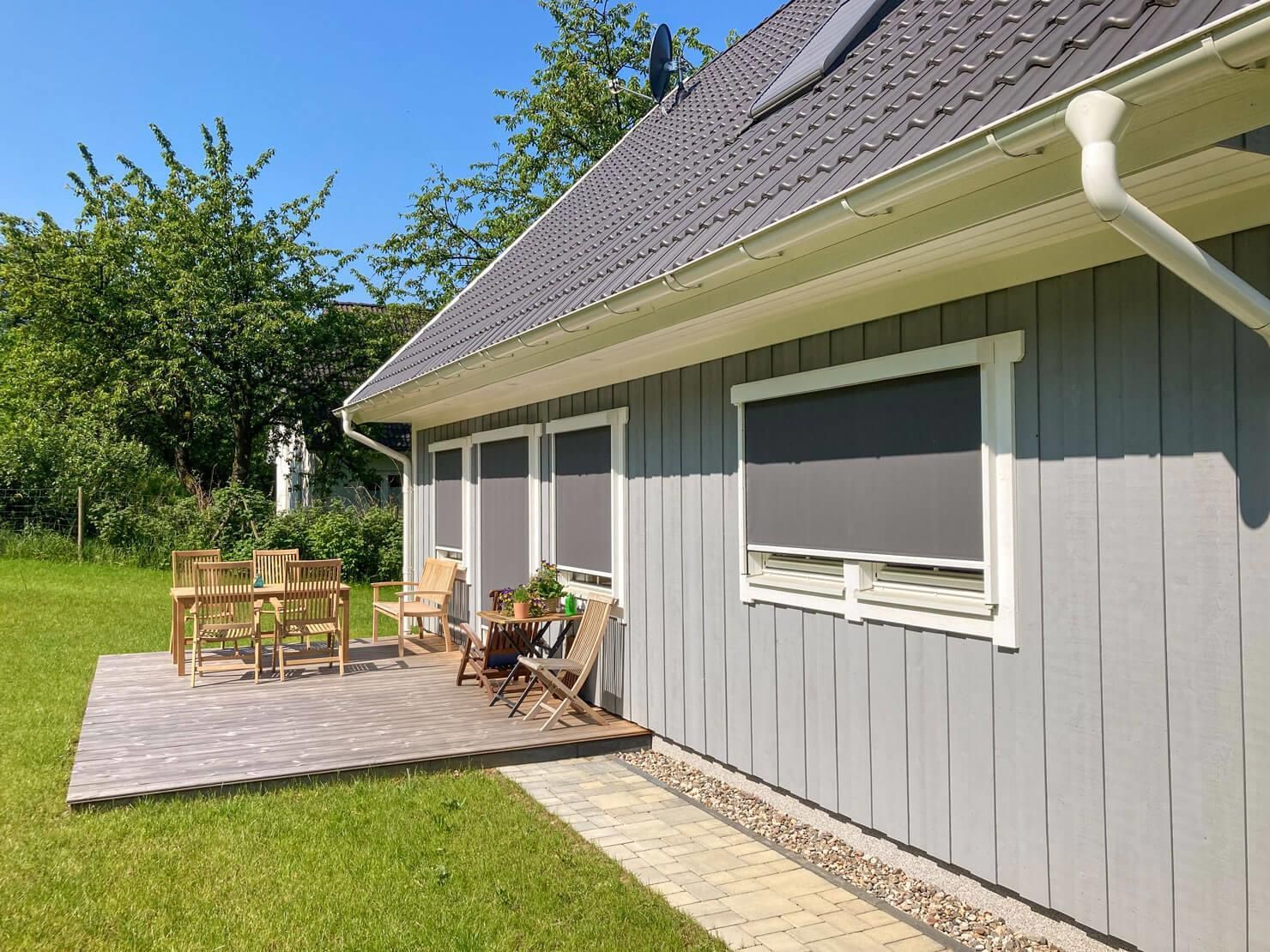 Graues Haus mit Sitzgelegenheiten im Freien auf einer gemauerten Terrasse, in einem grasbewachsenen Hof unter blauem Himmel.