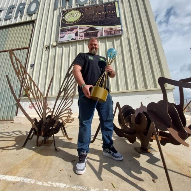 Un hombre con un trofeo se encuentra frente a una escultura metálica de un insecto. Exterior de un negocio.