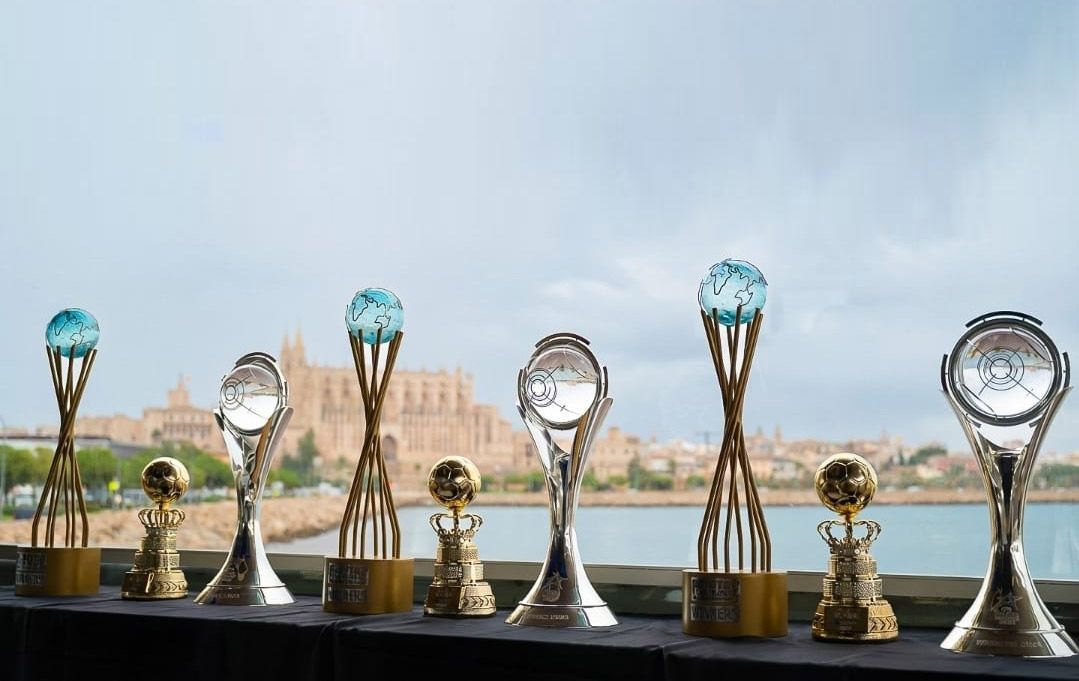 Trofeos sobre una mesa con una ciudad y agua al fondo bajo un cielo nublado.
