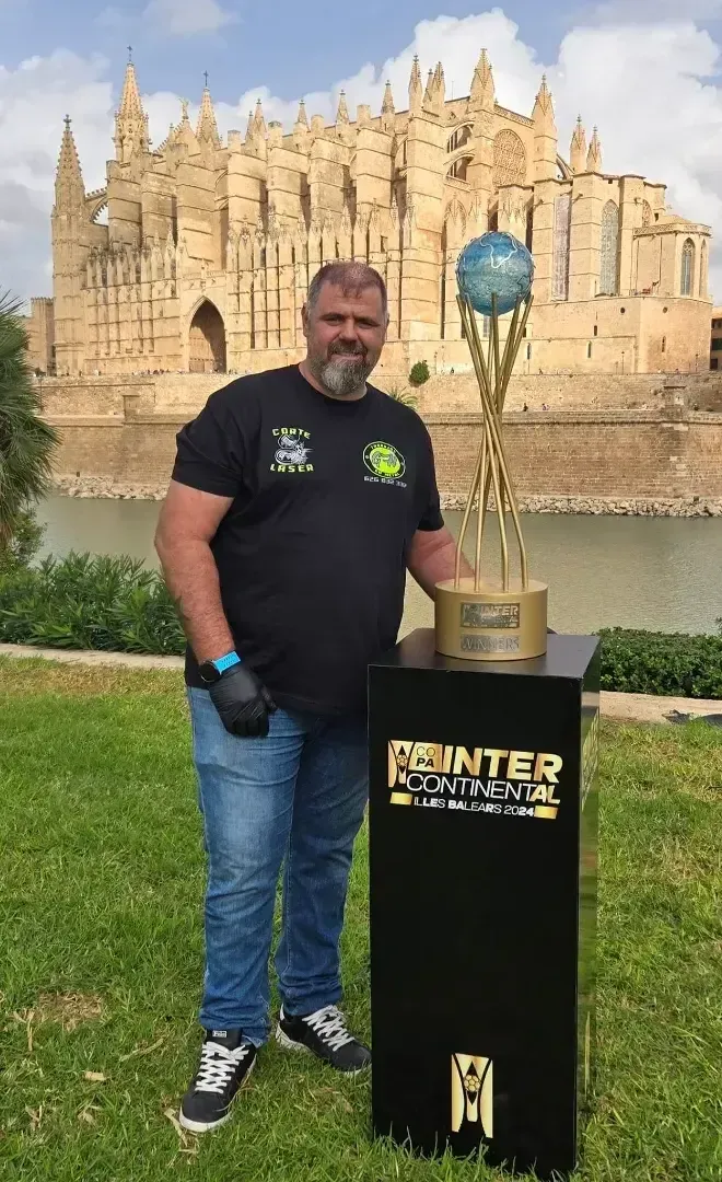 Hombre con un trofeo frente a una iglesia, posando para una foto. Camisa negra y vaqueros. En el expositor se lee