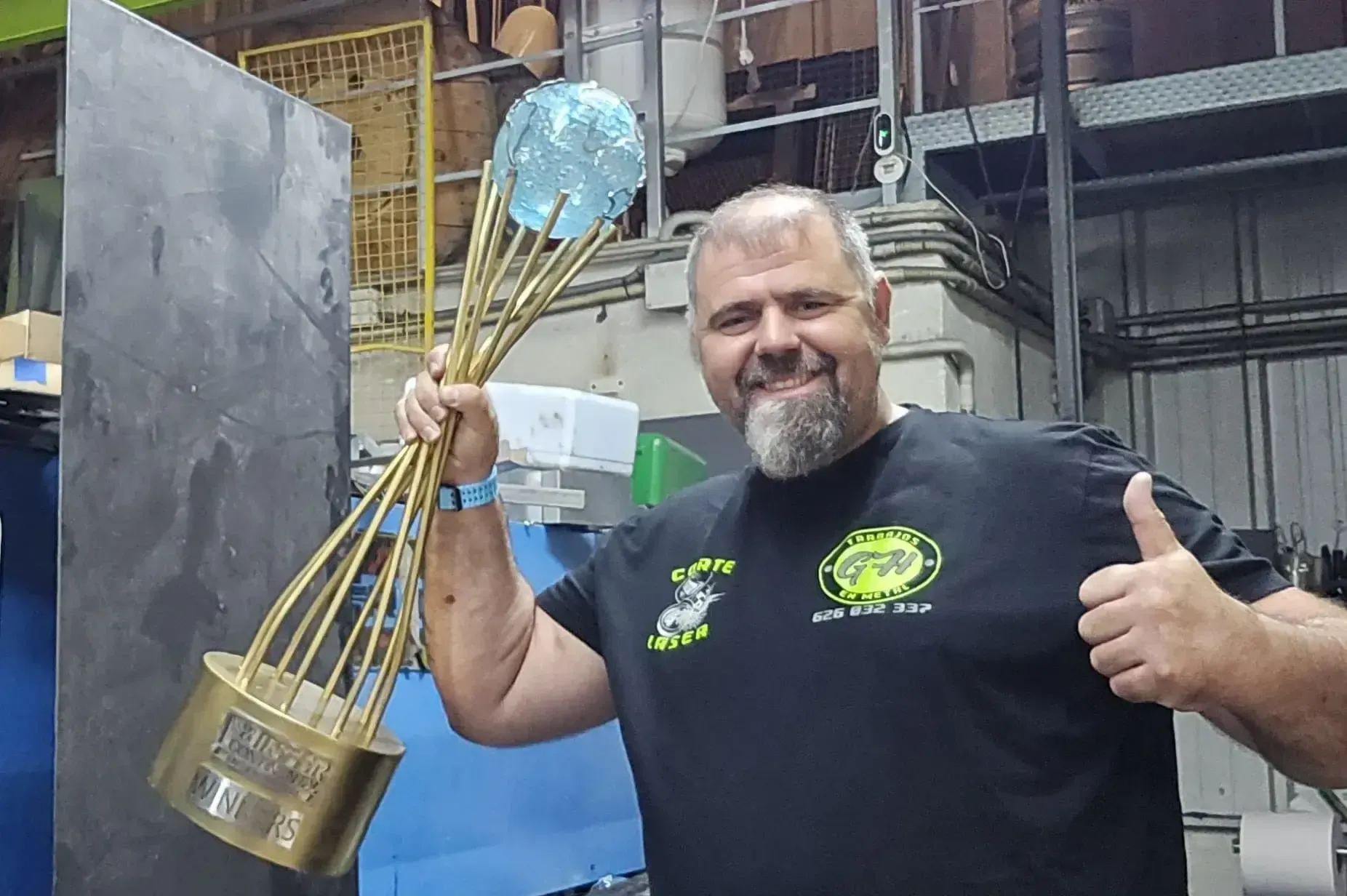 Hombre con un gran trofeo dorado, pulgares arriba. Está en un taller, sonriendo.