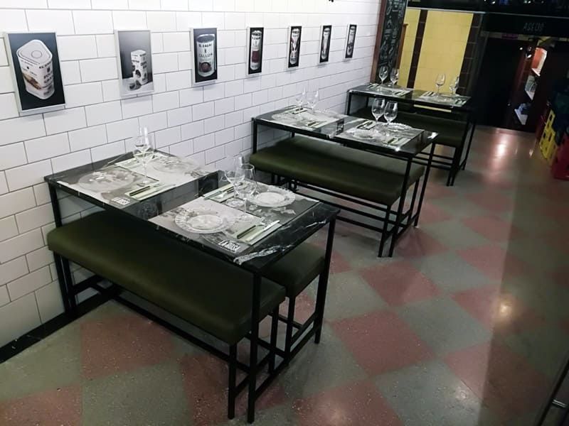 Zona de estar del restaurante con mesas negras, bancos verdes y suelo de cuadros. Una pared de ladrillo blanco exhibe obras de arte enmarcadas.