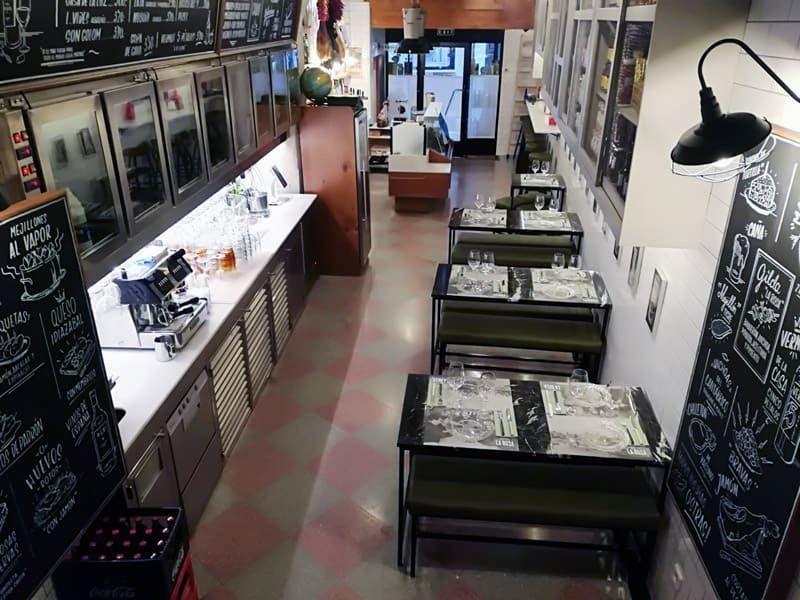 Interior de un restaurante con mesas, mostrador y pizarras. Suelo de baldosas rojas y blancas.