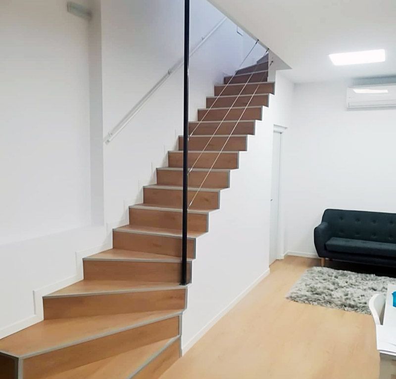 Escalera de madera en una habitación de paredes blancas con pasamanos metálico negro. Se ven un sofá y una alfombra.