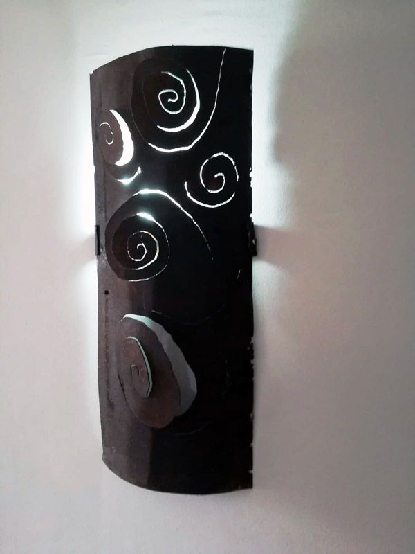 Aplique de pared negro con recortes en espiral, que emite luz sobre una pared blanca.